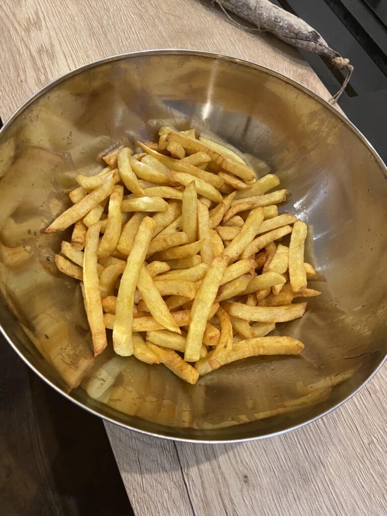 Frites croustillantes dans bol métallique, repas savoureux Frites dorées et chaudes servies dans un grand bol métallique brillant, posé sur une table en bois clair. Un saucisson sec repose à côté.