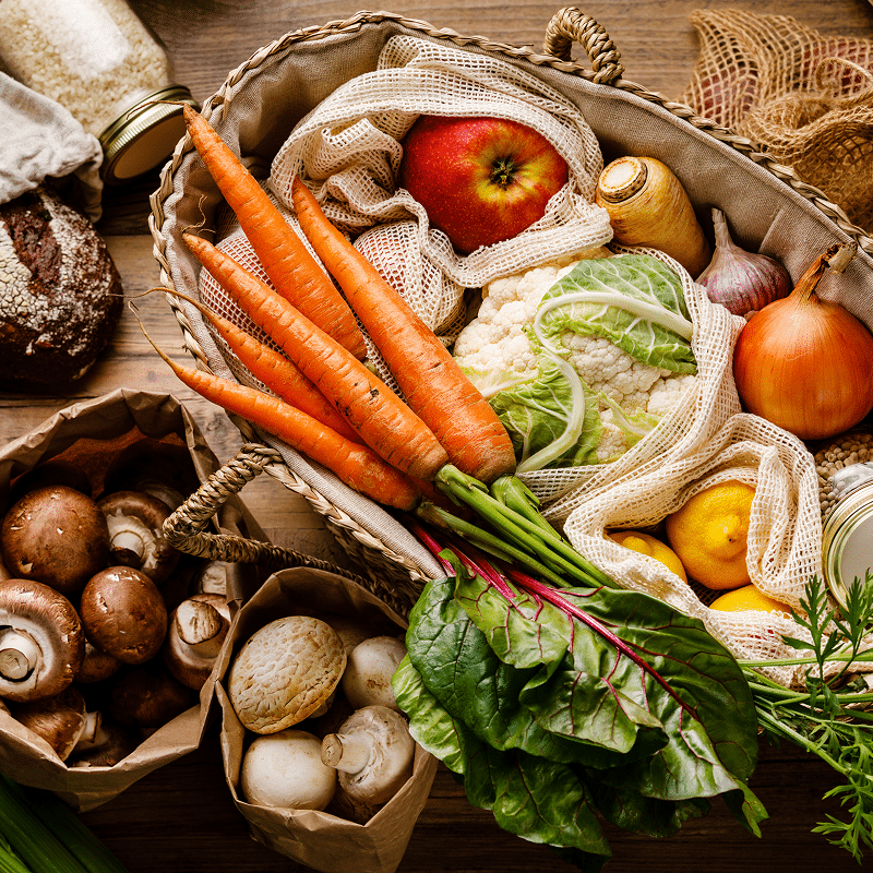 Gros plan d'un panier rempli de légumes frais : carottes, chou-fleur, betteraves, oignons, citrons, et champignons dans des sacs en papier.