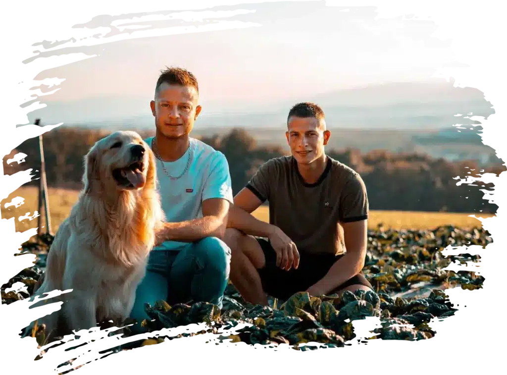 Deux hommes souriants et un Golden Retriever dans un champ de récolte au coucher du soleil, avec un paysage vallonné en arrière-plan.